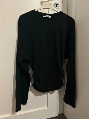 Svart långärmad topp NewYorker - En enkel svart långärmad topp från NewYorker med rund halsringning och utsvängda ärmar, den har även en ”scrunched up” look vid midjan, den sitter tight. Tillverkad i mjukt bomullsmaterial som känns skönt mot huden. Perfekt att styla med jeans eller kjol för en clean look. Tröjan är i storlek M.