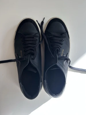 Svarta Axel Arigato sneakers - Svarta sneakers från Axel Arigato med lågt skaft, svart läder och mockadetaljer. Sidan pryds av en broderad fågel och guldfärgad logga.