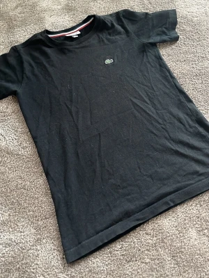 Svart t-shirt från Lacoste junior - Klassisk svart t-shirt från Lacoste med rund hals och den ikoniska gröna krokodilloggan broderad på bröstet. Tillverkad i mjuk bomull för en skön känsla. Perfekt till jeans eller shorts för en clean och enkel look. För barn