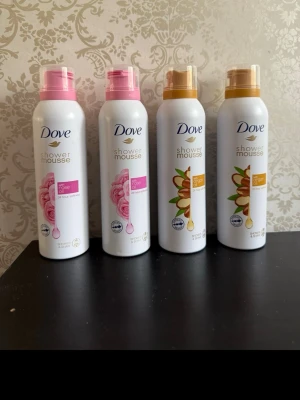 Dove Shower Mousse 4-pack - Fyra flaskor Dove Shower Mousse i olika dofter: två med ros och två med arganolja. Flaskorna är vita med rosa eller guldiga detaljer och har praktiska trycklock. Perfekt för dig som vill ha en lyxig duschupplevelse med mjukgörande mousse.