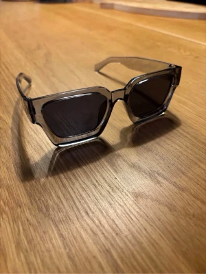 Genomskinliga fyrkantiga solglasögon - Säljer ett par stilrena solglasögon med transparent grå plastbåge och mörka, fyrkantiga linser. Glasögonen har en modern och edgy look som passar perfekt till alla outfits. Märke och storlek är okänt.