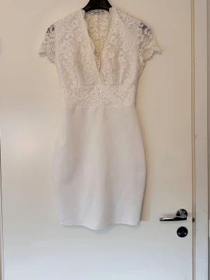Vit spetsklänning med v-ringning/studentklänning - Kort vit klänning med djup v-ringning och överdel i  dubbeltyg framtill med spets och genomskinlig baktill. Klänningen har korta ärmar och figurnära passform och är stretchig. Kjoldelen är slät och enkel medan överdelen har fina spetsdetaljer. Perfekt för dig som tar studenten eller att ha med utomlands