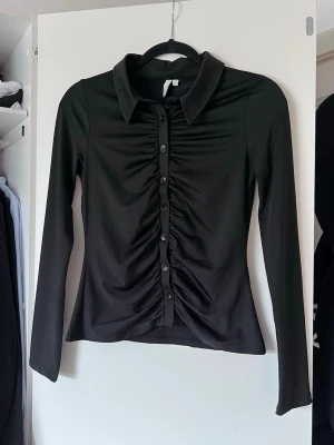 Y2K Svart långärmad topp med knappar från Nelly, stl S - Snygg svart topp med långa ärmar, knappar och krage. Materialet är stretchigt och följsamt, perfekt till många olika looks. 