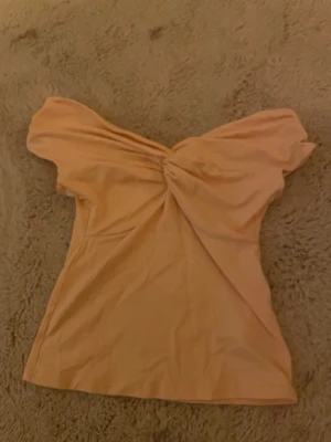 Persikofärgad offshoulder topp med twist - Trendig persikofärgad offshoulder topp med twistad detalj framtill. Toppen har en smickrande passform och är tillverkad i ett mjukt, stretchigt material som känns bekvämt mot huden. Perfekt för dig som vill ha en stilren men ändå unik look.