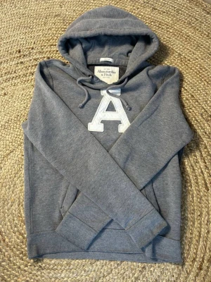 Grå hoodie från Abercrombie & Fitch - Snygg grå hoodie från Abercrombie & Fitch med stor vit broderad bokstav 'A' på bröstet. Tröjan har huva med dragsko och en klassisk känguruficka framtill. Perfekt för dig som gillar en avslappnad och cool stil. Den är i storlek m och i mycket bra skick hör av er vid frågor eller funderingar priset går att diskutera!!