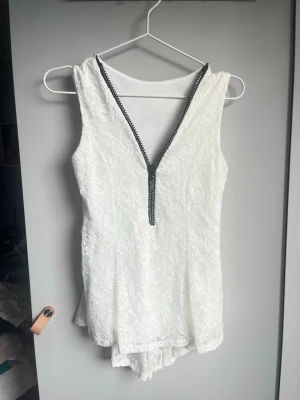 Vit spetslinne med v-ringad dragkedja - Säljer ett vitt ärmlöst spetslinne med v-ringning och dekorativ svart dragkedja framtill. Toppen har ett lätt utställd nederdel och är täckt av ett blommigt spetsmönster. Perfekt för dig som gillar romantiska detaljer och vill ha något extra till outfiten.Dragkedjan har diamanter på sig, om intresserad kan ni fråga om fler bilder.