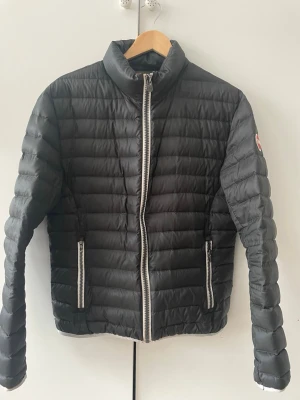 Svart pufferjacka från Colmar - Snygg svart pufferjacka från Colmar med quiltad design och hög krage. Jackan har vita dragkedjor framtill och på fickorna samt Colmar-logga på ärmen. Tillverkad i lätt och smidigt material som håller värmen under kalla dagar.