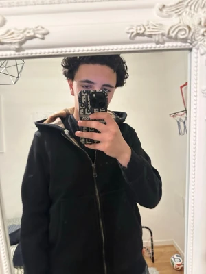 Svart hoodie från Burberry med dragkedja - Säljer en svart hoodie från Burberry med dragkedja framtill och klassiskt rutigt foder i huvan. Hoodien har två fickor fram och en stilren design som passar perfekt till streetwear. Materialet känns mjukt och bekvämt, och huvan ger extra stilpoäng.