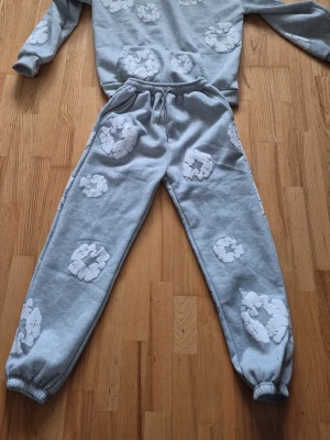 Grått mjukisset  Denimtears - Säljer ett grått mjukisset med sweatshirt och matchande byxor. Båda plaggen har vita nallemönster tryckta över hela ytan. Tröjan har en känguruficka framtill och byxorna har resår i midjan och vid bensluten. Materialet är mjukt och skönt, perfekt för chill dagar hemma.