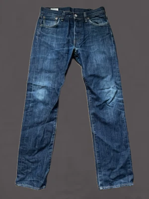 Levi's 501 - Klassiska blå jeans från Levi's med rak passform och fem fickor. Jeansen har en snygg tvättad look, silverfärgade nitar och knapp, samt Levi's ikoniska läderpatch bak i midjan. Tillverkade i slitstarkt denimtyg som passar till många olika stilar. Tyvär trasiga i skrevet men är relativt lättfixat