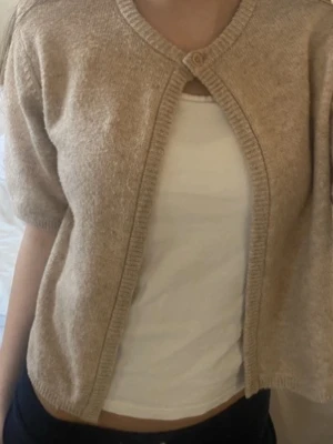 Beige kortärmad kofta med knapp - Säljer en beige, kortärmad kofta med rund hals och en knapp framtill. Koftan har ribbade kanter och är gjord i ett mjukt stickat material. Perfekt att slänga över ett linne för en chill och enkel look.