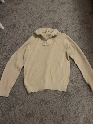 Beige stickad tröja med halvzip - Snygg beige stickad tröja med halv dragkedja och hög krage. Tröjan har lång ärm och ribbade muddar vid ärmslut och nederkant. Perfekt för lager på lager och enkel att matcha med olika outfits.