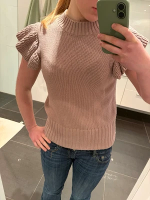 Rosa stickad topp med volangärm - Säljer en rosa stickad topp med ribbad halsringning och nederkant. Toppen har korta volangärmar som ger en söt och trendig look. Perfekt att styla med jeans eller kjol för en chill vibe. Tröjan har nästan aldrig använts. Den är från Lindex, specifikt Holly & White.