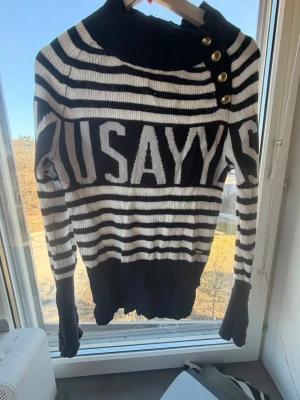 Randig svartvit stickad tröja - Stickad tröja i svart och vitt med breda ränder och texten 'USAYYAS' över bröstet. Hög krage med tre dekorativa guldknappar på axeln. Långa ärmar och ribbade muddar. Tröjan har en avslappnad passform och är perfekt för en trendig streetwear-look.