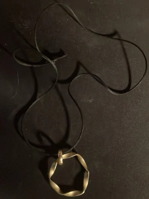 Svart halsband med vriden metallring - Unikt halsband med svart snöre och en vriden, sexkantig ring i guldfärgad metall som hänge. Snygg och enkel design som sticker ut med sin geometriska form. Perfekt accessoar för dig som gillar minimalistisk stil.OBS! Materialet är okänt, kan ej garantera att det är rostfritt.