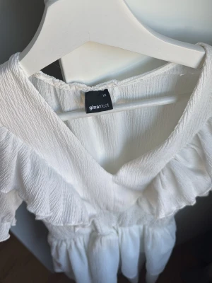 Vit volangklänning från Gina Tricot - Supersöt vit kortklänning från Gina Tricot med v-ringning, volangdetaljer längs axlar och bröst samt smockad midja. Klänningen är ärmlös och har en luftig, flowy passform som ger en romantisk vibe.