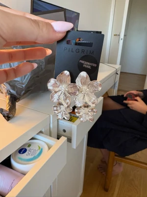 Silvriga blomörhängen från Pilgrim - Stora statementörhängen i silverfärg från Pilgrim, formade som blommor med detaljerade blad och en liten pärldetalj i mitten. Örhängena har en exklusiv look och är märkta med 'Freshwater Pearl'. Perfekta för dig som gillar att sticka ut.