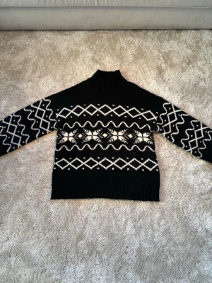 Svartvit stickad tröja Gina Tricot - Stickad oversized svart tröja från Gina Tricot med vitt grafiskt mönster över bröstet och ärmarna. Tröjan har hög krage och ribbstickade muddar vid hals, ärmslut och nederkant. Perfekt för kalla dagar och ger en trendig look.