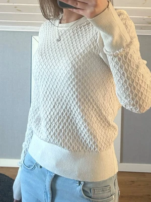 Vit stickad tröja med strukturmönster - Säljer en superfin vit stickad tröja med snygg struktur och rund hals. Tröjan har långa ärmar och breda ribbade muddar vid ärmslut och nederkant. Perfekt till jeans för en clean och fräsch look. Väl använd och är lite smått nopprig men syns inte på håll, en liten fläck på ena ärmen men märks inte av. 