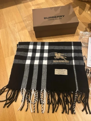 Svart-vit rutig halsduk från Burberry - Snygg halsduk från Burberry i klassiskt svart och vitt rutmönster med broderad logga och fransar i ändarna. Tillverkad i mjukt material som känns lyxigt och trendigt. Perfekt accessoar för att lyfta din outfit.