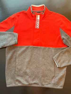 Hugo Boss tröja - Snygg tröja från BOSS i rött och grått med hög krage och dragkedja. Stora BOSS-loggan framtill på kragen. Tröjan har långa ärmar, kontrastfärgade muddar och två sidofickor. Perfekt för en avslappnad men stilren look.