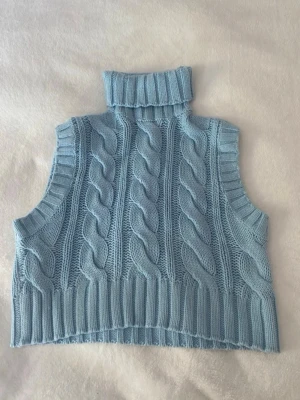 Ljusblå stickad poloväst - Trendig ljusblå stickad väst med polokrage och breda kabelstickade detaljer framtill. Västen är croppad och har ribbade kanter vid ärmhål och nederkant. Perfekt att styla över en skjorta eller t-shirt för en cool lager-på-lager-look.