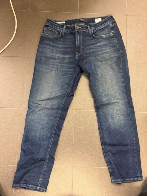 Blå raka jeans från Jack and Jones  - Säljer ett par klassiska blå jeans från Jack and Jones med raka ben och fem fickor. Jeansen har snygga slitningar framtill och bakfickor med diskret söm. Tillverkade i bomull med en bekväm passform och knappgylf.