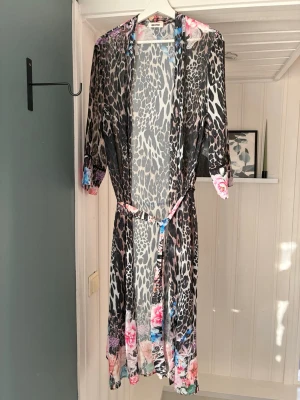 Leopardmönstrad  långkofta - Säljer en tunn kofta motsv med leopardmönster i svart, grått och vitt. Kanterna är dekorerade med färgglada blommor i rosa och blått. Koftan har trekvartsärmar och ett matchande knytband i midjan. 