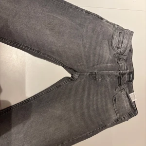 Grå jeans från jack&jones - Säljer ett par grå jeans med klassisk femficksmodell och raka ben. Jeansen har snygga slitningar och kontrastsömmar. Perfekta för en avslappnad och trendig look.
