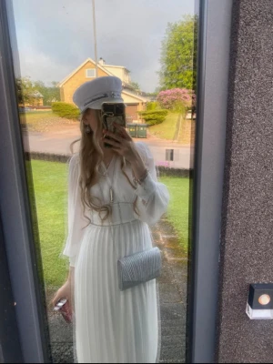 Studentklänning  - Elegant vit långklänning med plisserad kjol och transparenta långärmar. Klänningen har v-ringning och markerad midja, vilket ger en klassisk och stilren look. Perfekt till student eller sommarens högtider.