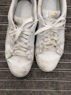 Vita Puma sneakers - Snygga vita sneakers från Puma med guldfärgad logga på plösen. Skorna har klassisk låg profil, vita snören och ovandel i slätt läder. Perfekta för en clean och sportig look. Diskreta sömmar och tidlös design gör dem lätta att matcha till olika outfits.