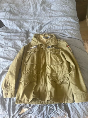 Olivgrön parkajacka med patchar - Säljer en olivgrön parkajacka med flera fickor och coola patchar på bröstet och ärmen. Jackan har krage, knappar och snörning i midjan för en snygg passform. Perfekt för dig som gillar militärinspirerad stil.