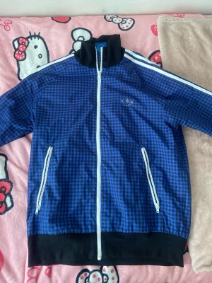 Blårutig Adidas jacka med dragkedja - Säljer en blå och svart rutig tunn jacka från Adidas med vita detaljer och dragkedja framtill. Tröjan har två fickor med vita dragkedjor, svart krage och vita ränder på axlarna. Adidas-logga på bröstet. Perfekt för en sportig och avslappnad stil.