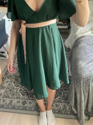 Grön omlottklänning med bälte - Snygg grön omlottklänning med v-ringning och korta ärmar. Klänningen har ett beige knytbälte i midjan som ger en fin siluett. Tyget är mjukt och faller snyggt, perfekt för varmare dagar.