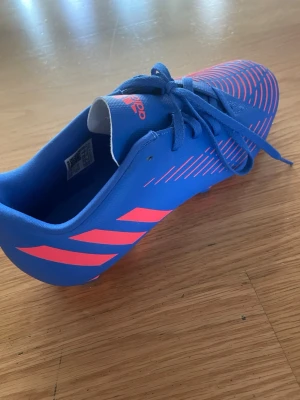 Adidas Predator Edge.4 blå fotbollsskor - Snygg blå fotbollssko från Adidas Predator Edge.4 med rosa detaljer och klassiska ränder på sidan. Skon har snörning och är tillverkad i syntetmaterial för bra grepp och kontroll på planen. Perfekt för dig som vill sticka ut på matchen. Har använt dem en eller två gånger så i helt nytt skick