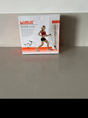 Lineup Door Gym träningsband - Säljer ett Lineup Door Gym träningsband som fästs i dörren för mångsidig hemmaträning. Perfekt för styrketräning och stretching. Kommer i originalförpackning med handtag och fästen. Färg: svart och orange.