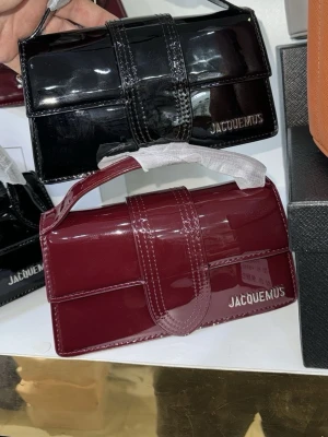 Jacquemus handväska  - Snygg svart handväska från Jacquemus i glansigt lackmaterial. Väskan har ett stilrent, rektangulärt format med bred rem över locket och ett kort handtag. Jacquemus-loggan i silver syns på framsidan. Perfekt accessoar för att lyfta din outfit.