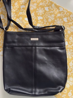 Svart axelväska från Don Donna - Stilren svart axelväska från Don Donna i slätt konstläder. Väskan har justerbar axelrem och en enkel, fyrkantig form med diskret metallogo framtill. Perfekt för dig som vill ha en klassisk och enkel väska till vardagslooken.