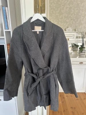 Grå ullkappa från H&M med skärp - Grå kappa från H&M i ullblandning. Modellen har bred krage, knytskärp i midjan och raka ärmar. 