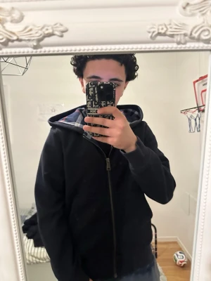 Mörkblå hoodie med rutigt foder - Säljer en mörkblå hoodie från Burberry med dragkedja och stor huva. Insidan är fodrad med ett mjukt rutigt tyg i blått, vitt och rött. Perfekt för dig som vill ha en enkel men snygg hoodie med lite extra detaljer. Passar till det mesta och är riktigt bekväm.