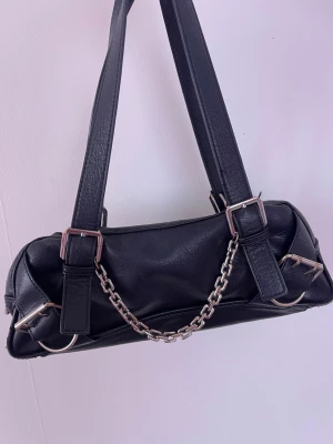 Svart axelväska med kedjedetalj - Snygg svart handväska i avlång modell med dubbla axelband och silverfärgade kedjedetaljer framtill. Väskan har dragkedja upptill och coola metalldetaljer på sidorna. Perfekt accessoar för en edgy look. Dragsjedjan kan gå ur från plats som du drar för snabbt! Den lite rosting men inget som märks