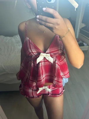 Rött rutigt pyjamas-set med rosetter - Säljer ett sött pyjamas-set i röd- och vitrutigt mönster. Setet består av ett linne med smala axelband och djup urringning samt matchande shorts. Båda delarna har dekorativa vita rosetter framtill och volangkant. Perfekt för mysiga kvällar hemma. Från Gina tricot. Hela settet är i storlek xs