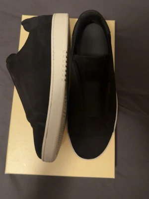 Svarta Axel Arigato sneakers - Stilrena svarta slip-on sneakers från Axel Arigato med vit sula och minimalistisk design. Skorna har diskret guldfärgad logga på sidan och är tillverkade i mocka. Perfekta för dig som gillar clean och modern stil. Nästan helt ny och bara använd 2-3 gånger. Original pris är 3000 kronor. Möts upp i Göteborg
