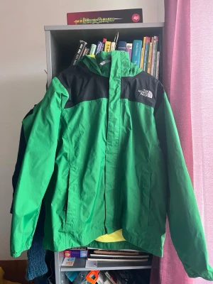 Grön vindjacka för barn från The North Face, 134 - Snygg vind-/regnjacka storlek 10 år.The Märke: North Face. Grön med svarta axelpartier och huva. Jackan har dragkedja framtill, två sidofickor och logga på bröstet. Perfekt för blåsiga dagar och outdoor-äventyr. Tillverkad i slitstarkt syntetmaterial. Mycket fint skick, inget slitage.