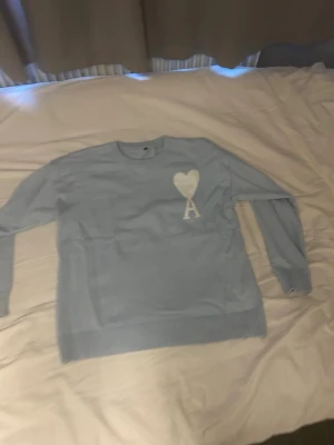 Ljusblå stickad tröja från Ami Paris - Ljusblå stickad tröja från Ami Paris med vitt hjärta och bokstaven 'A' broderat på bröstet. Tröjan har rund halsringning, ribbade muddar och lång ärm. Perfekt för dig som gillar stilrena detaljer.