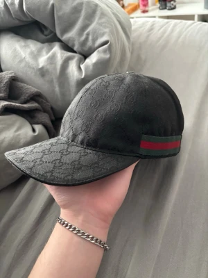Svart Gucci keps med GG-mönster - Säljer en svart keps från Gucci med klassiskt GG-mönster i tyget. Kepsen har en böjd skärm och en grön-röd rand på sidan som ger den en ikonisk look. Justerbar rem baktill för perfekt passform. Materialet känns lyxigt och stilrent.