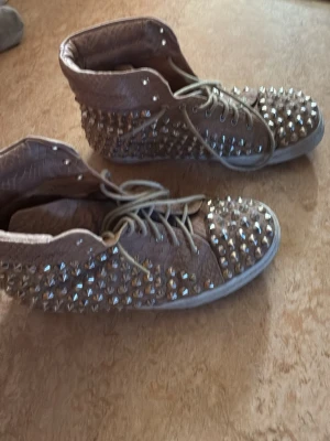 Lanvin liknade Beige sneakers med nitar och ormmönster - Unika beige sneakers med högt skaft, täckta av silvriga nitar och med ett coolt ormmönstrat material. Skorna har vita snören och en klassisk gummisula. Perfekta för dig som vill sticka ut och addera lite edge till din outfit.( inte lanvin)