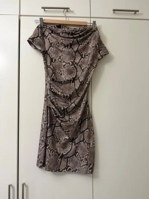 Kort klänning med ormmönster - Säljer en kort, figurnära klänning med ormmönster i beige och bruna toner. Klänningen har korta ärmar och en mjuk, stretchig passform. Perfekt för dig som vill sticka ut med ett djurmönster. Ingen synlig dragkedja eller knappar.