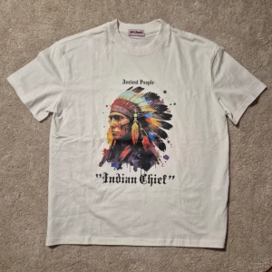 Palm Angels - Säljer en vit t-shirt från Palm Angels HELT NY!!med ett stort färgglatt tryck av en fjäderprydd huvudbonad och texten 'Ancient People' samt 'Indian Chief' på framsidan. T-shirten har rund hals och korta ärmar. Perfekt för dig som gillar unika prints.