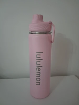 Rosa vattenflaska från Lululemon - Snygg rosa vattenflaska från Lululemon med praktiskt skruvlock och bärhandtag. Flaskan är tillverkad i rostfritt stål och har en matt finish. Perfekt för träning, skola eller vardagsbruk. Lululemon-loggan syns tydligt på sidan.